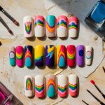 A variety of colorful false nail designs are arranged in rows. Some feature rainbow patterns, while others have abstract art. The scene is surrounded by nail polish bottles and brushes on a textured beige surface.