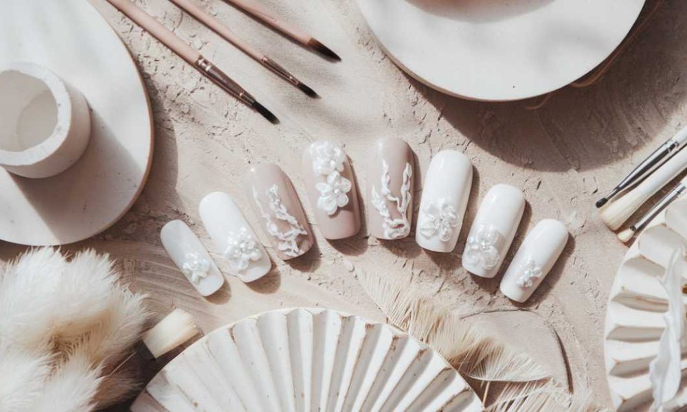 A set of beige and white press-on nails with intricate floral designs. The display is surrounded by art brushes, white decorative feathers, and ceramic plates on a textured, sandy surface.