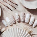 A set of beige and white press-on nails with intricate floral designs. The display is surrounded by art brushes, white decorative feathers, and ceramic plates on a textured, sandy surface.