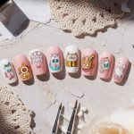 A set of artificial nails with cartoon designs including a strawberry, donut, face, ice cream, sneaker, bee, macaron, and hot dog. They are placed on a textured surface surrounded by brushes, crochet pieces, and decorative shells.