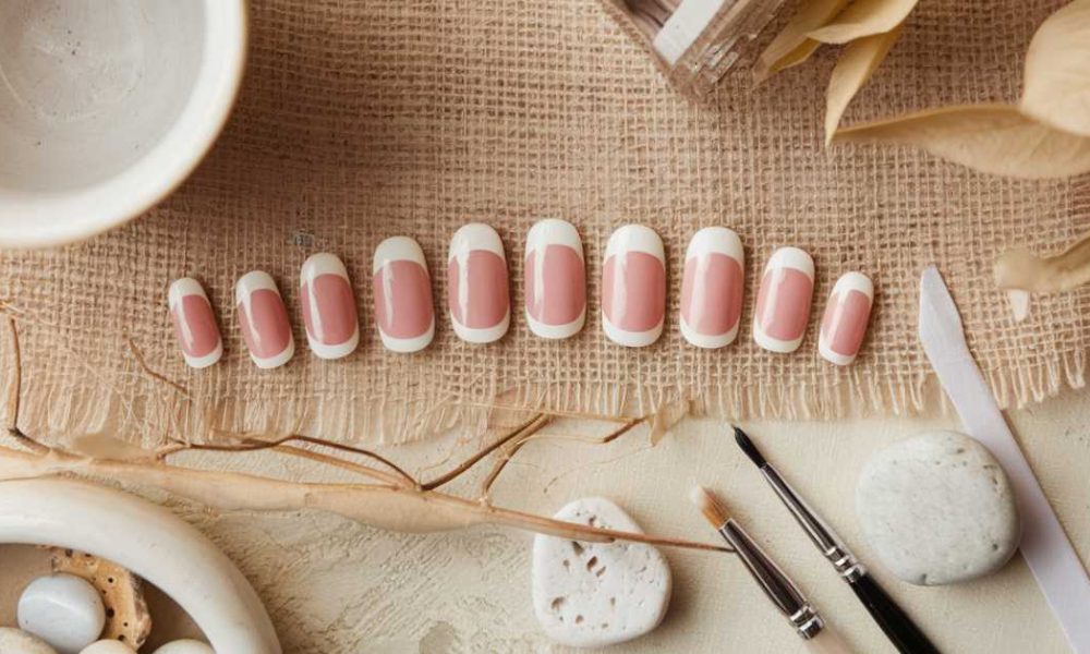 A set of artificial nails with a pink base and white tips displayed on a burlap cloth. Nearby are nail tools, white stones, a ceramic dish, and dried leaves, creating a rustic, natural aesthetic.