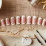 A set of artificial nails with a pink base and white tips displayed on a burlap cloth. Nearby are nail tools, white stones, a ceramic dish, and dried leaves, creating a rustic, natural aesthetic.