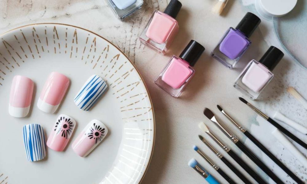 A set of fake nails with pink, blue, and floral designs displayed on a plate. Nearby are bottles of nail polish in pink, blue, and purple shades, along with various nail art brushes on a white and pastel background.