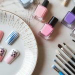 A set of fake nails with pink, blue, and floral designs displayed on a plate. Nearby are bottles of nail polish in pink, blue, and purple shades, along with various nail art brushes on a white and pastel background.