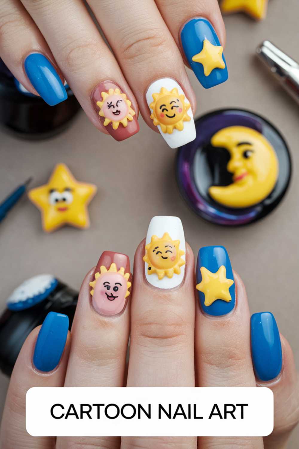 Close-up of hands showcasing Cartoon Nail Art Designs. Nails are painted blue and white, adorned with 3D sun, moon, star, and cloud designs. The background is dotted with cute cartoon figurines of stars and a moon. Text on image: CARTOON NAIL ART.