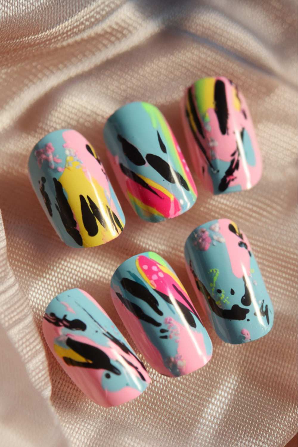Close-up of cute nail art showcasing pastel-colored press-on nails with abstract designs in pink, yellow, blue, and black on a shimmery, light fabric background.