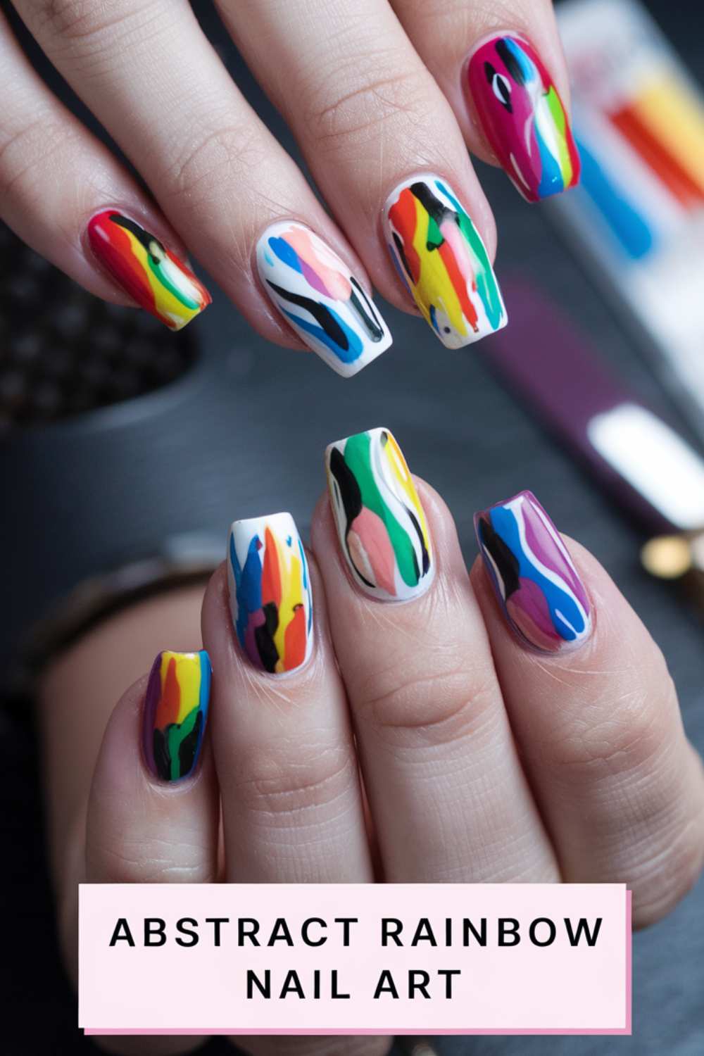 Close-up of hands showcasing mesmerizing rainbow nail art. Each nail boasts a unique, vibrant design bursting with colors like red, blue, green, yellow, and pink. The text Abstract Rainbow Nail Art elegantly graces the bottom.