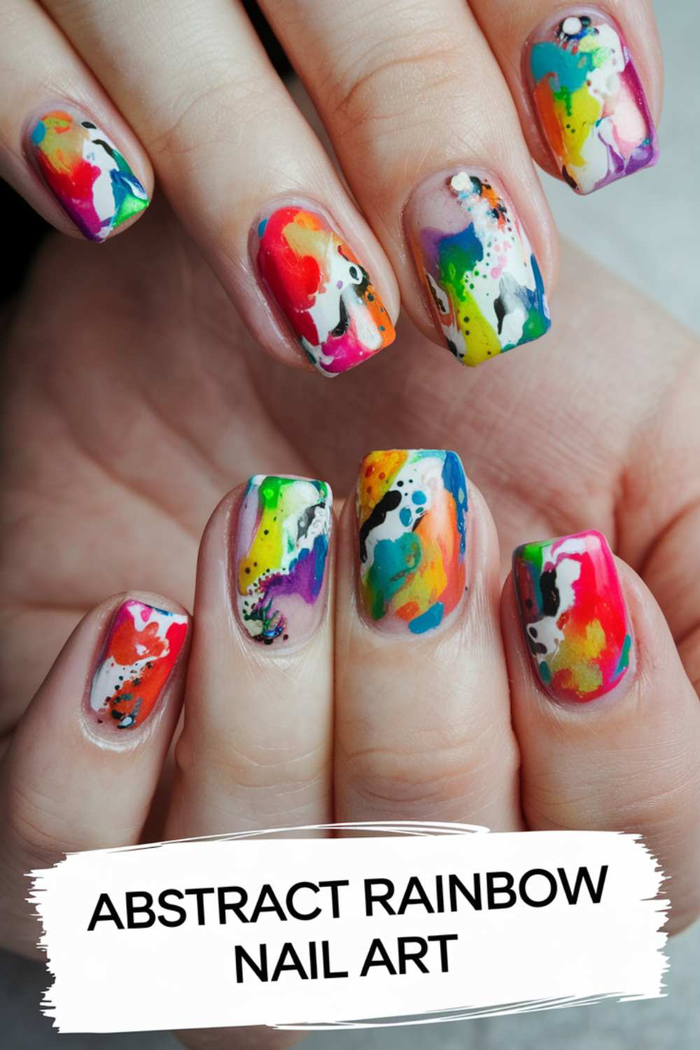 Close-up of hands with abstract rainbow nail art showcasing vibrant splashes of red, blue, yellow, green, and purple. The modern design features white accents and black dots. Text at the bottom reads Abstract Rainbow Nail Art, celebrating a bold expression of personal style.