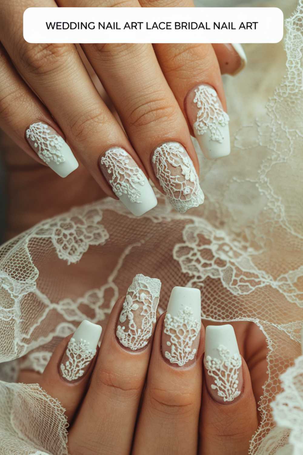 Close-up of a hand with elegant white lace bridal nail art on long nails. The intricate lace designs complement the lace fabric in the background, creating a cohesive wedding-themed image.