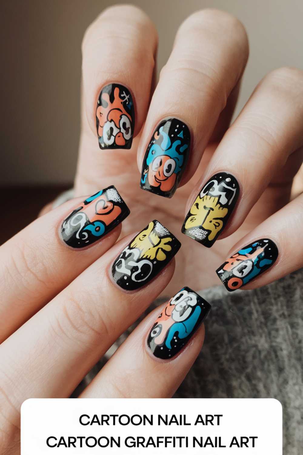 Close-up of a hand with nails featuring vibrant cartoon nail art designs on a black background. The colorful graffiti includes playful faces and abstract shapes. Text at the bottom reads Cartoon Nail Art Cartoon Graffiti Nail Art.