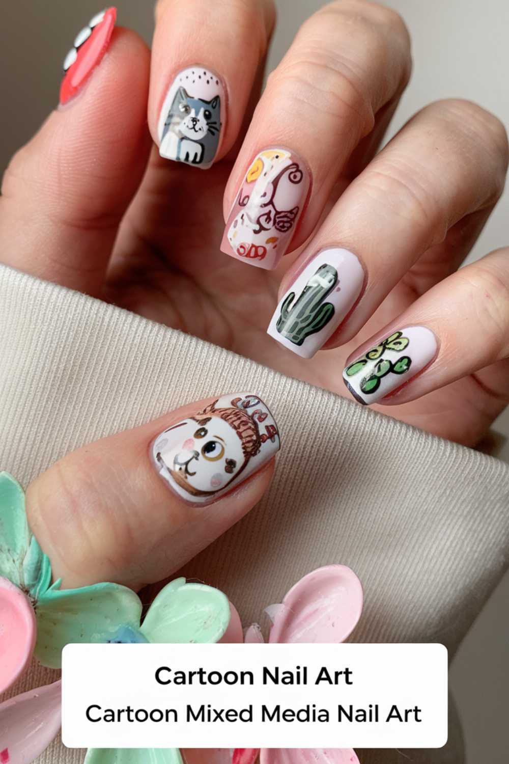 The hand showcases vibrant cartoon nail art designs, featuring a cat, llama, and cactus among playful images. These lively nails stand out against a cream sweater, while the thumb sports a colorful floral decoration.