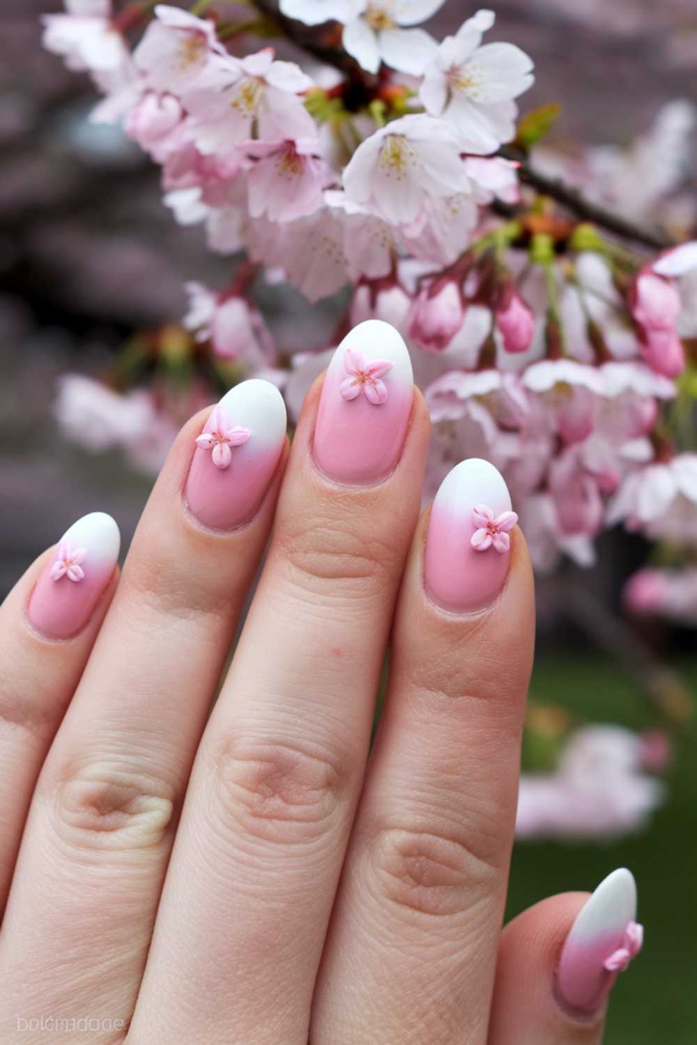 A hand with manicured nails featuring pink and white ombre design and 3D flower accents. The background shows blossoming pink cherry blossoms on branches.