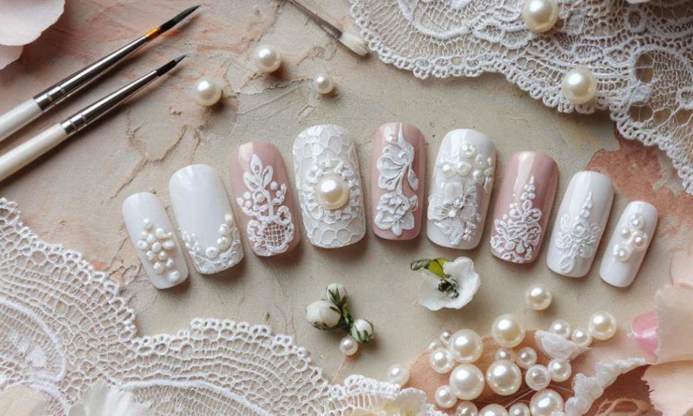 Nail art display with intricate lace and pearl designs on artificial nails. The nails feature white and blush tones, surrounded by small paintbrushes, lace, pearls, and delicate white flowers on a textured surface.