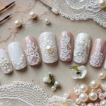 Nail art display with intricate lace and pearl designs on artificial nails. The nails feature white and blush tones, surrounded by small paintbrushes, lace, pearls, and delicate white flowers on a textured surface.