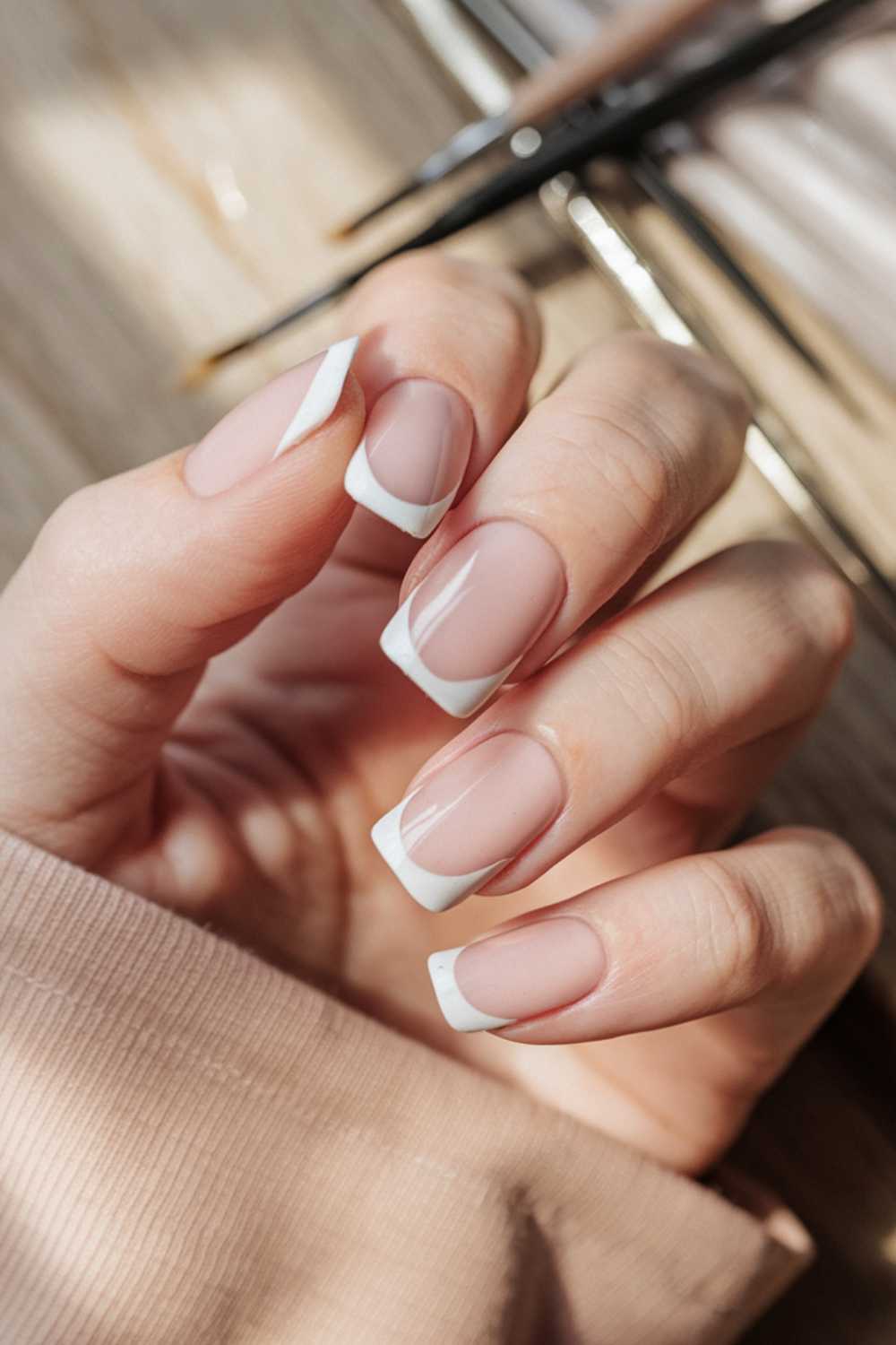 A hand with a classic French manicure, showcasing neatly shaped nails with pink bases and white tips. The hand rests on a wooden surface, and the person is wearing a beige long-sleeve top.