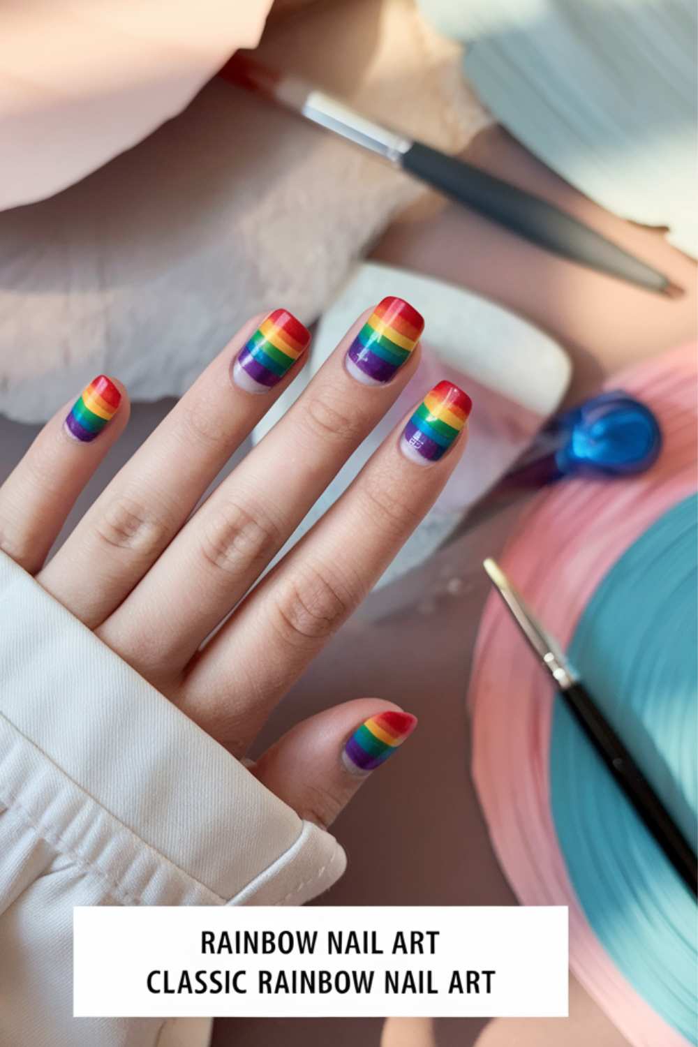 A hand with exquisite rainbow nail art on each fingernail rests gracefully on a white surface. Nearby, pastel blue and pink circular objects accompany a nail brush and a small blue heart-shaped object. A label proudly declares, Classic Rainbow Nail Art.