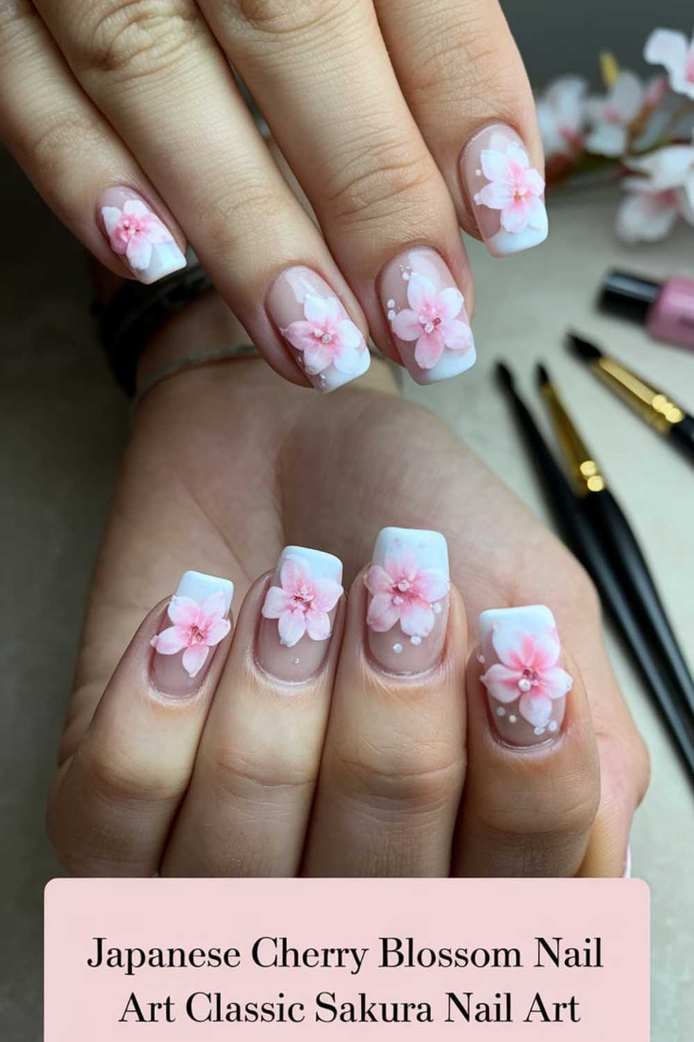 Close-up of hands with nails featuring cherry blossom art. The square-shaped nails are adorned with pink and white flowers, creating a classic Sakura look. Brushes and a nail polish bottle are visible in the background. Text reads: Japanese Cherry Blossom Nail Art Classic Sakura Nail Art.