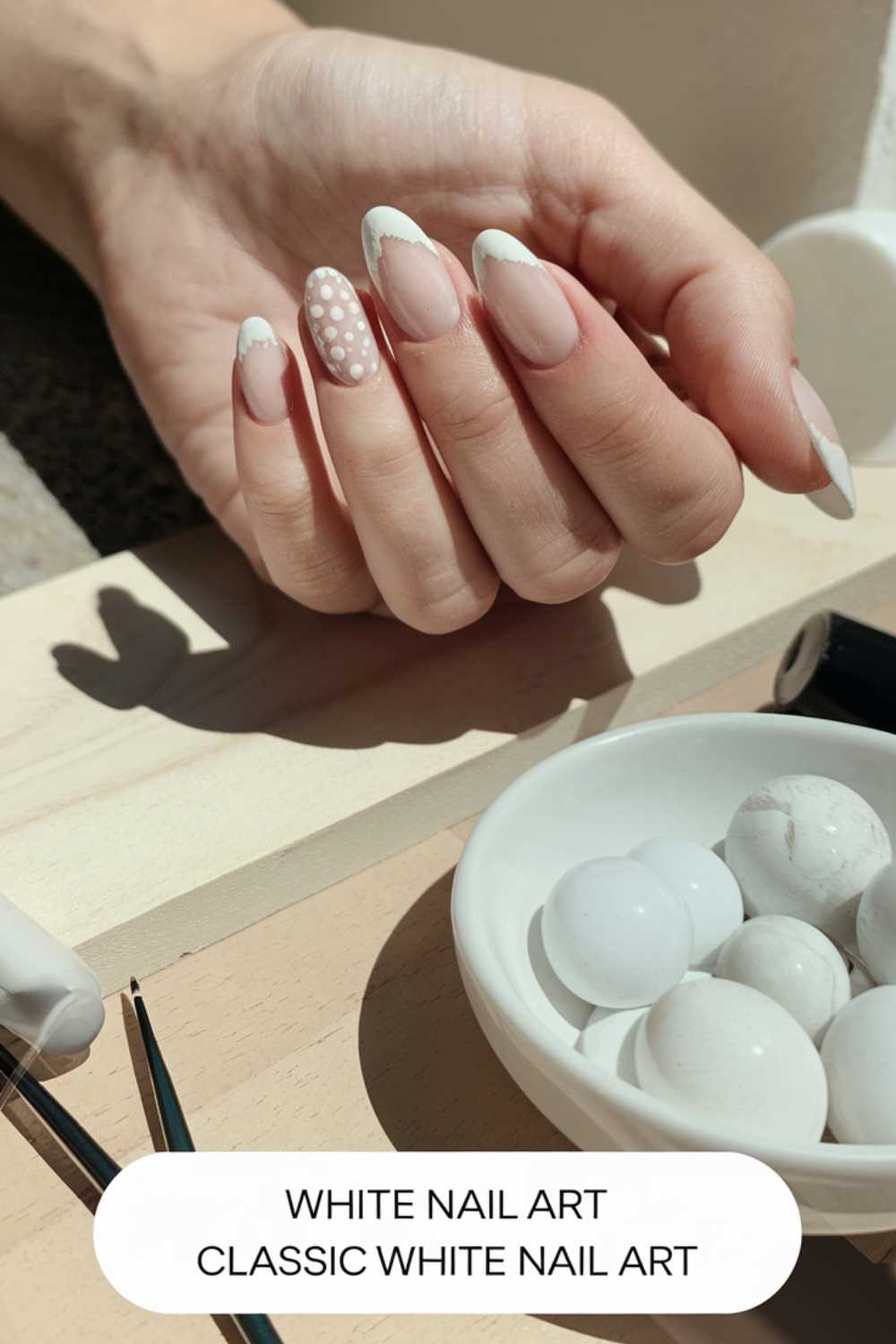 A hand shows a set of manicured nails with white French tips and one accent nail featuring polka dots. The background includes a wooden surface, nail art tools, and round marble-like objects. Text reads WHITE NAIL ART CLASSIC WHITE NAIL ART.