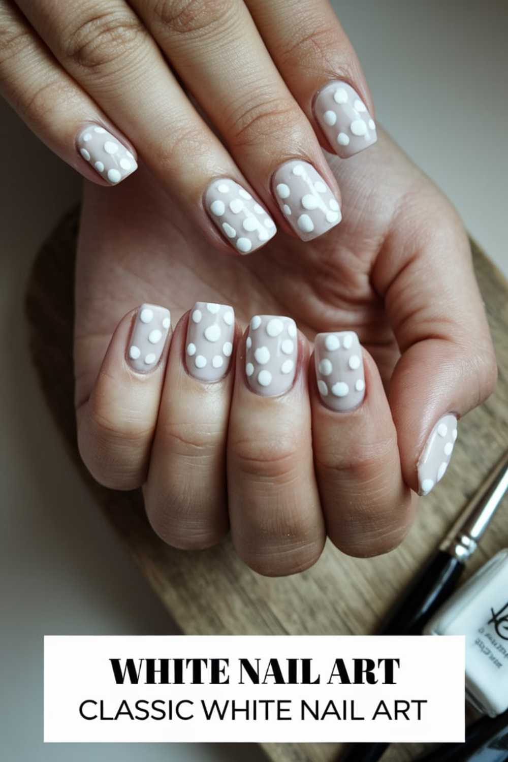 Hands with white nail art featuring small white dots on a neutral base. A text overlay reads WHITE NAIL ART and CLASSIC WHITE NAIL ART. A wooden surface and a nail polish bottle are visible in the background.