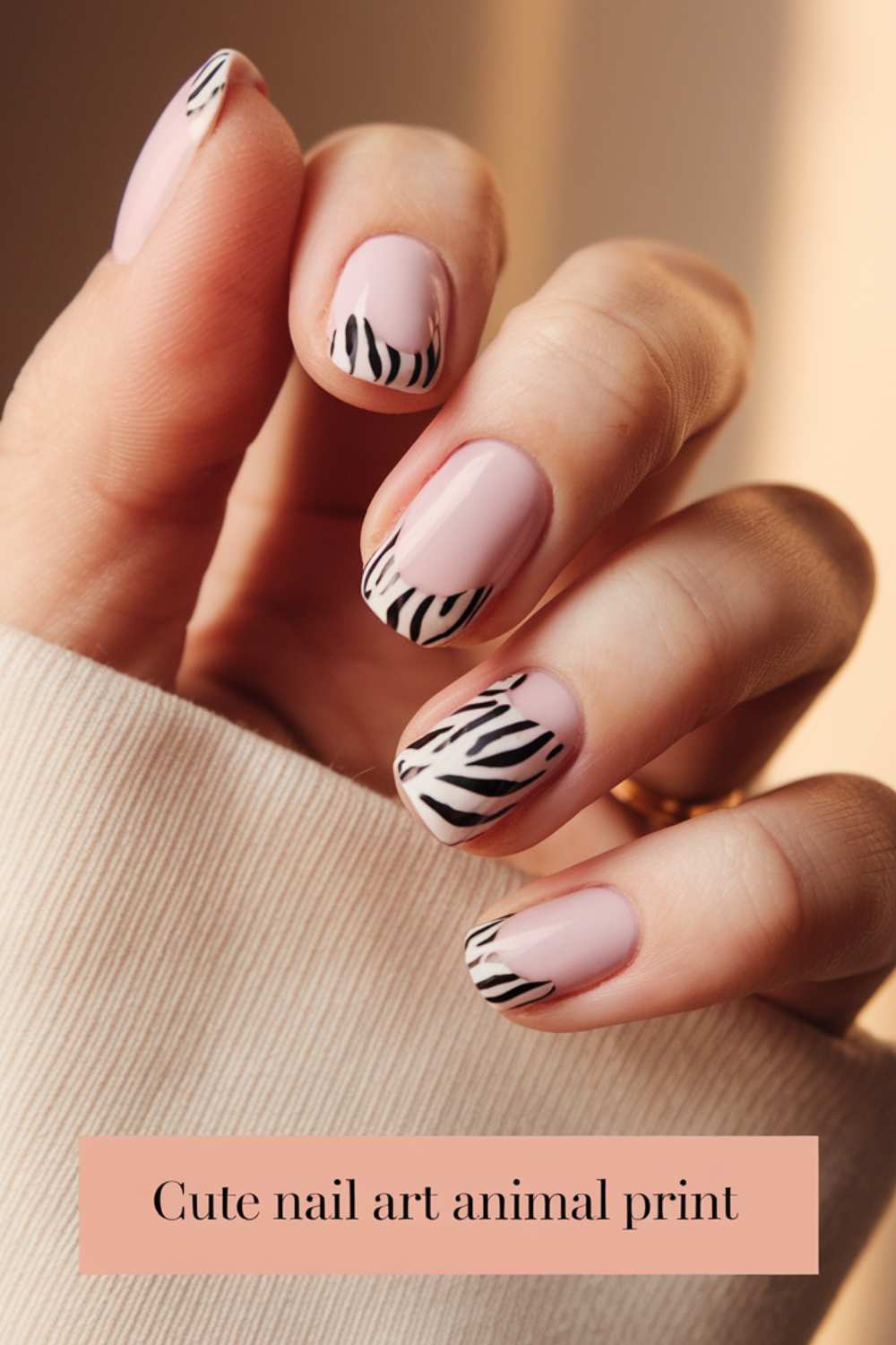 Close-up of a hand with nails painted in a light pink shade, featuring black and white zebra stripes on the tips. The hand is positioned gracefully, wearing a beige sleeve. Text at the bottom reads, Cute Nail Art showcasing chic animal print design.