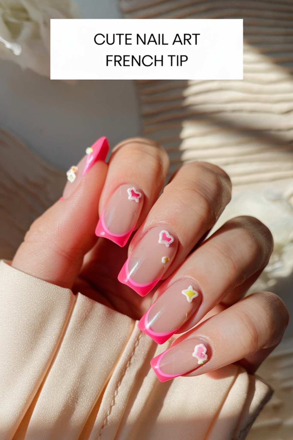 A hand with a French tip manicure showcases bright pink tips adorned with small, colorful charms like stars and hearts. Against a ribbed fabric background in sunlight, the caption reads, Cute Nail Art French Tip.