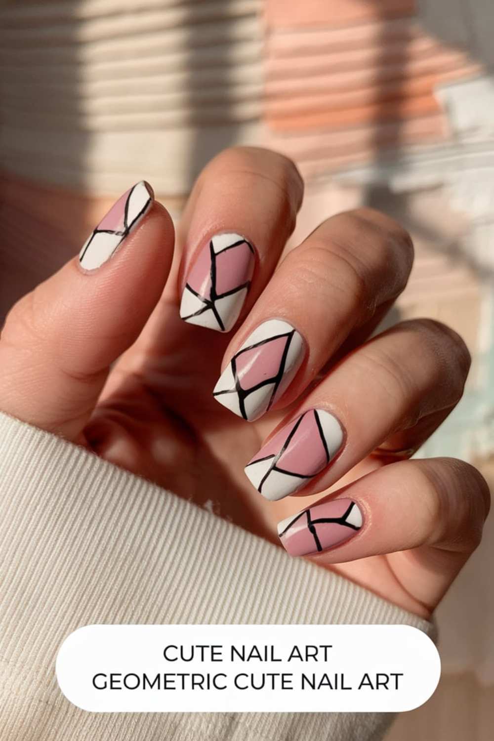 A hand flaunts cute geometric nail art with pink and white segments accented by bold black lines for a stained-glass look. The square-shaped nails, paired with a cozy white sweater, create an adorable style statement.
