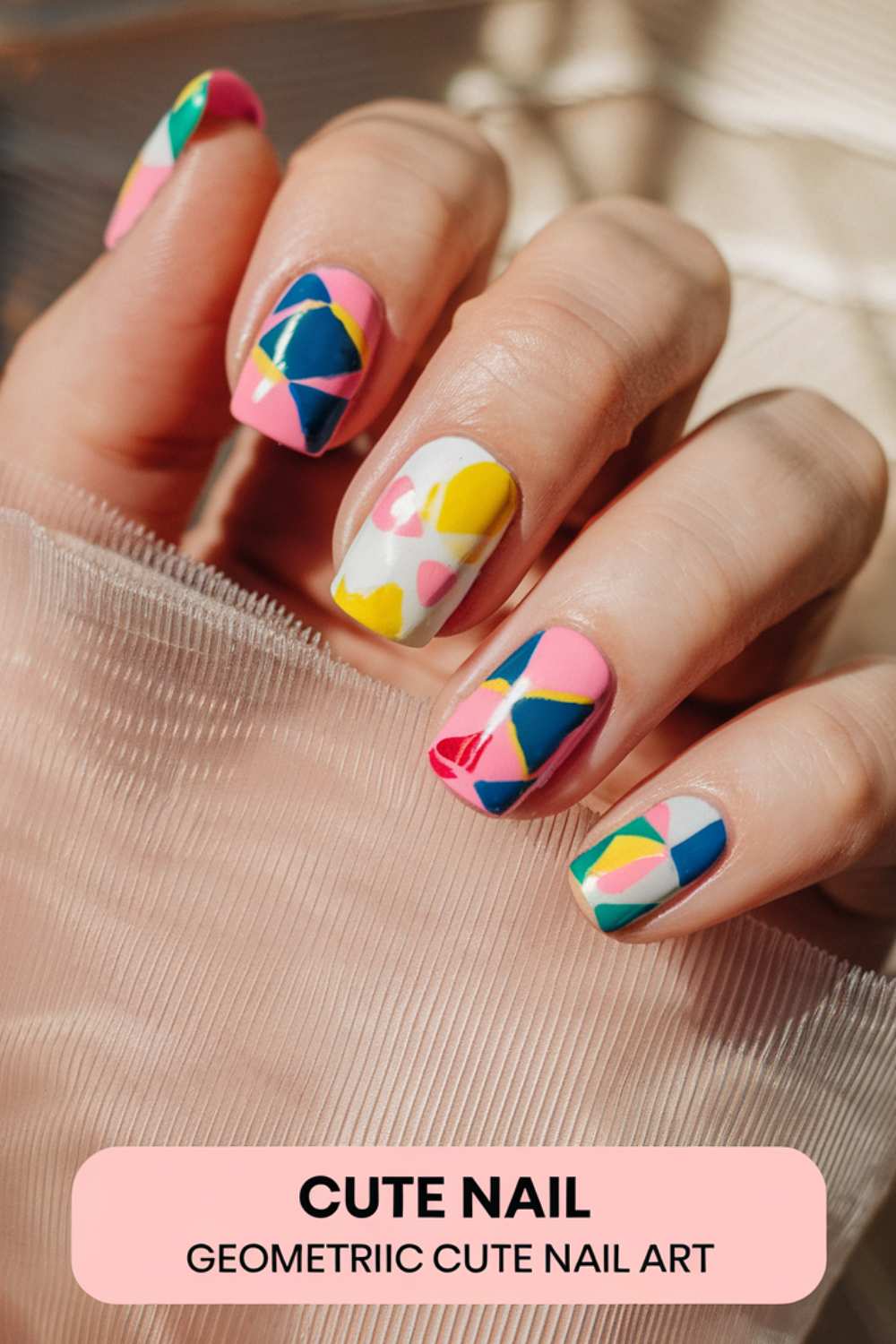 A hand showcases vibrant geometric nail art with pink, blue, yellow, and white shapes against a textured, translucent fabric. Text at the bottom reads: CUTE NAIL ART - GEOMETRIC CUTE NAIL ART.