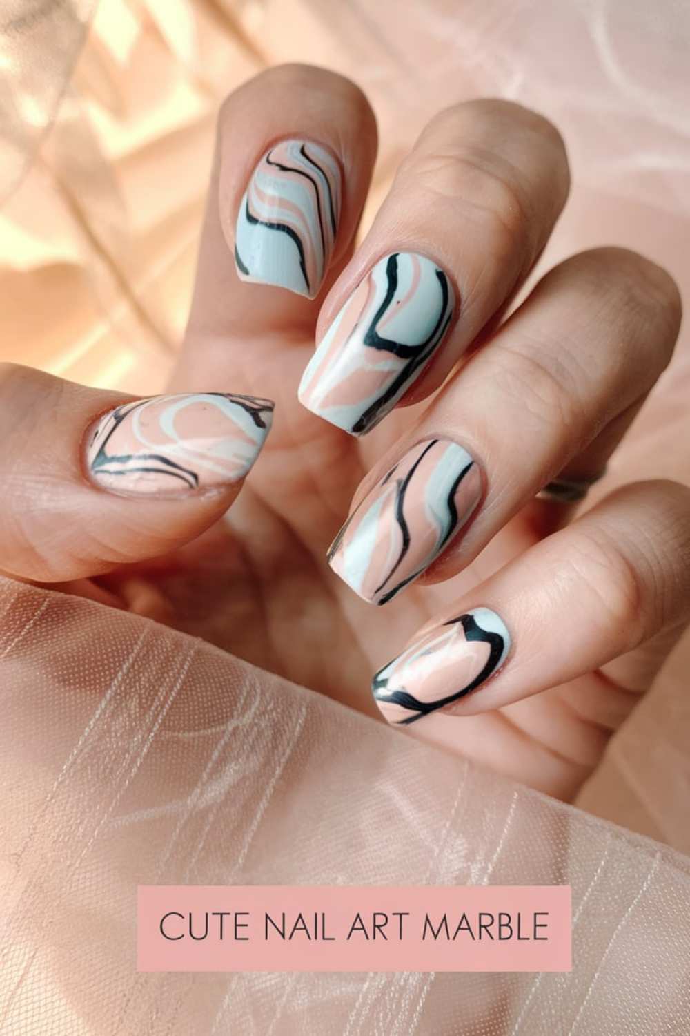 A hand with nails showcasing marble nail art in shades of pink, white, and black. The soft peachy background enhances the cute nail art design. Text at the bottom reads, Cute Nail Art Marble.