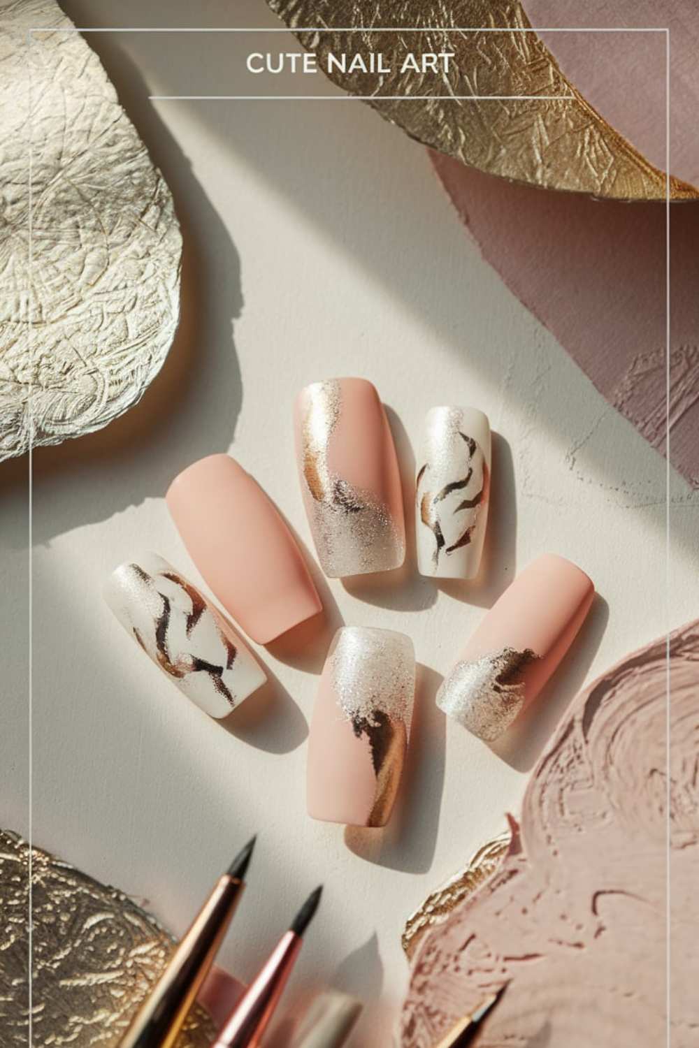 A set of six artificial nails displayed on a surface, featuring a matte peach color with marble and metallic gold accents. Artistic brushes and textured paper surround them, adding an elegant touch to this cute nail art.