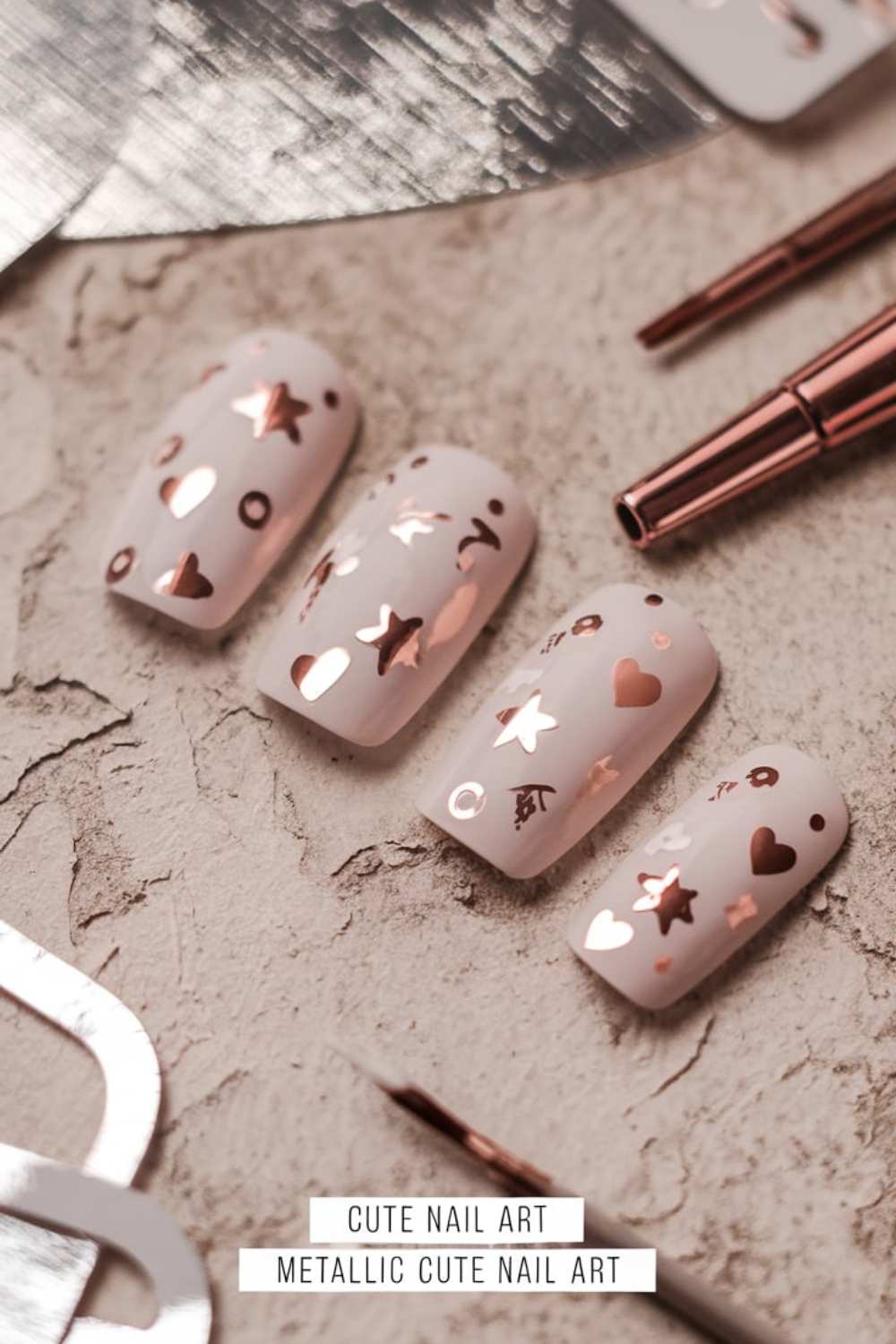 Four fingernail tips with light pink nail polish showcase cute nail art, featuring metallic designs of stars, hearts, and polka dots. These charming motifs are arranged on a textured surface with nail art tools nearby.
