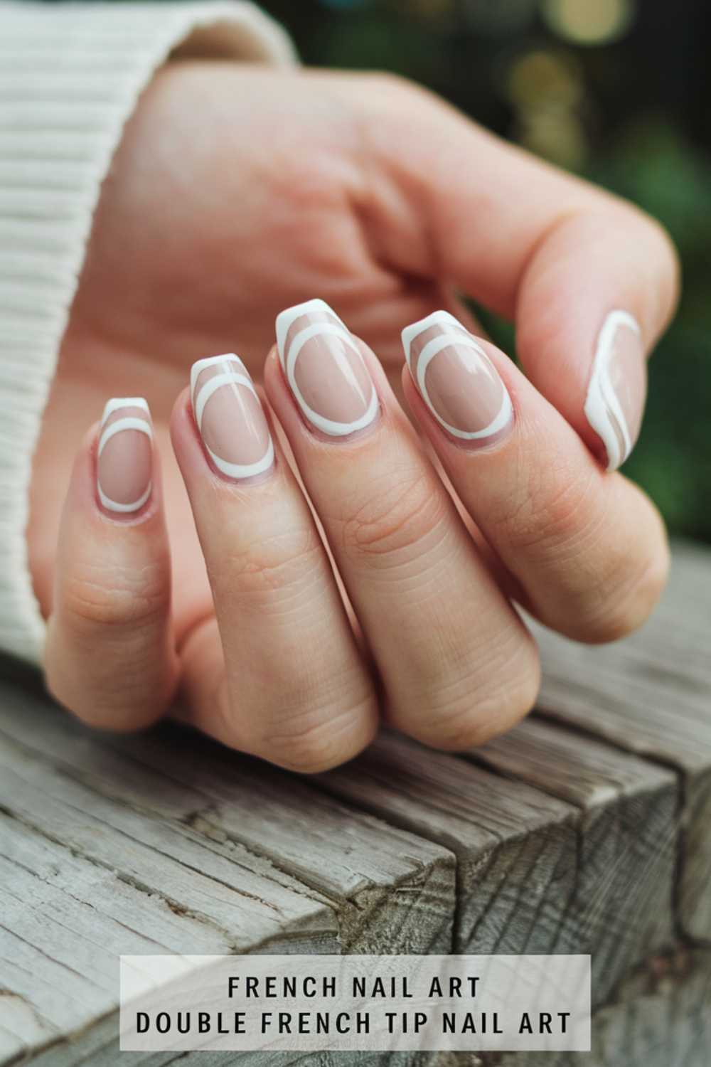 Close-up of a hand with an innovative French manicure featuring double white tips on each fingernail. The nails rest on a wooden surface. The person is wearing a cream sweater. Text at the bottom reads: “FRENCH NAIL ART DOUBLE FRENCH TIP NAIL ART.”.
