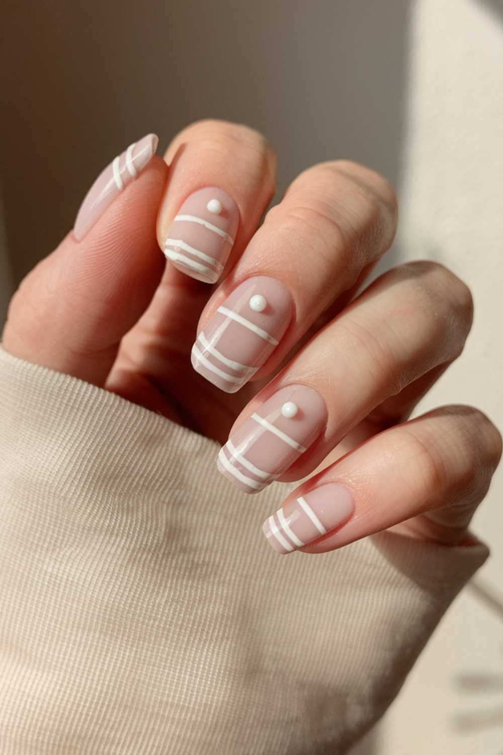 A hand with a light-colored sleeve showcases manicured nails featuring simple yet minimalist nail art. The nails have a nude base with white geometric lines and dots, creating an elegant, modern look.