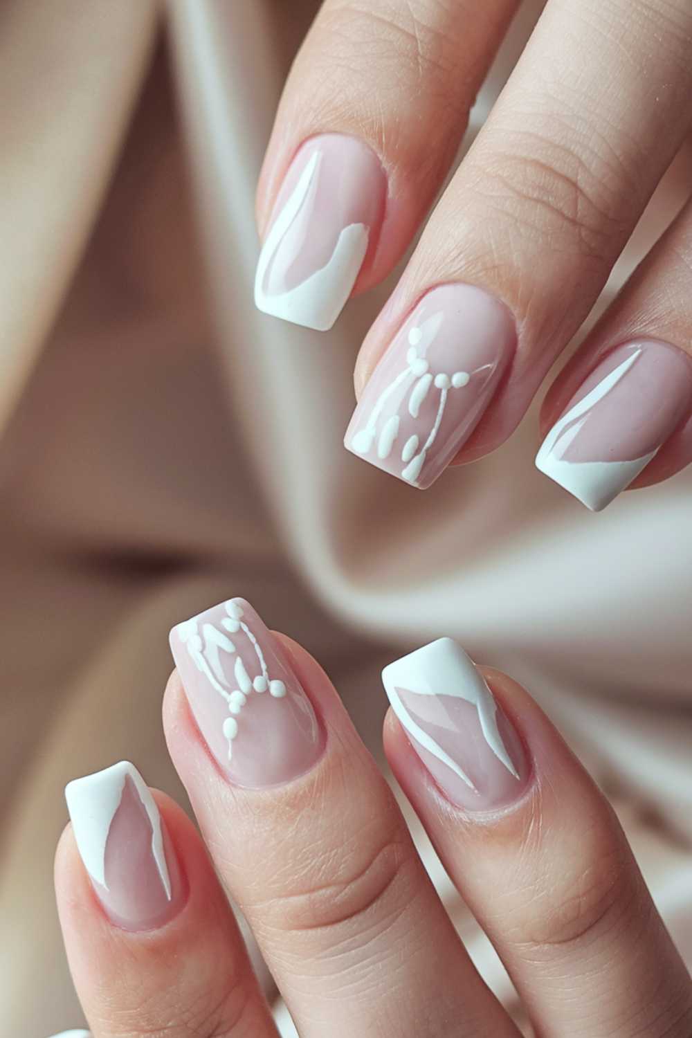Close-up of a hand with an elegant French manicure showcasing simple nail art. The nails feature white tips with glossy pink bases, while two nails boast intricate white designs resembling delicate patterns, set against a soft, beige fabric background.