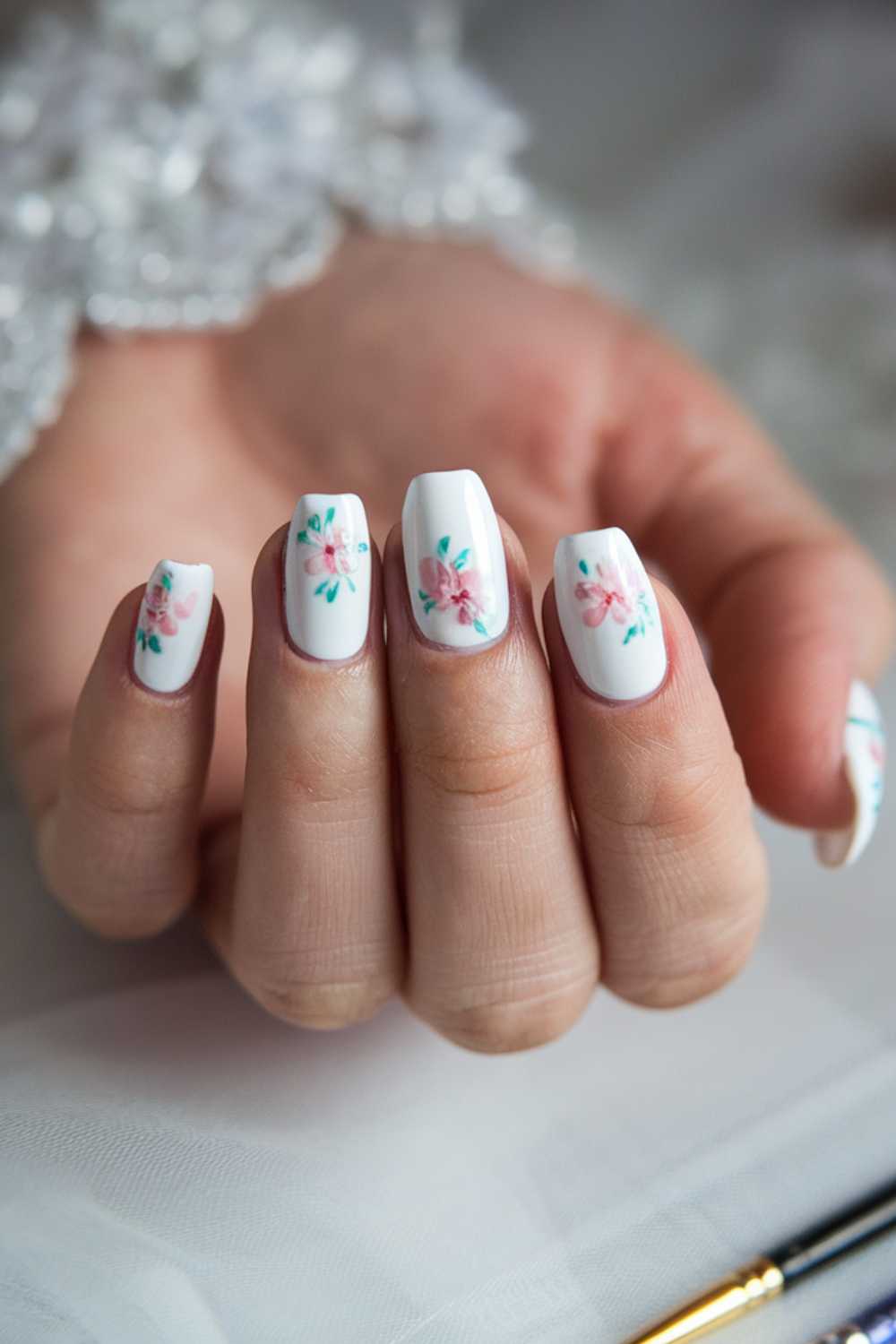 A hand with long, manicured nails painted white featuring delicate pink floral designs with green leaves. The fingers are slightly curled, and the background is soft and blurred.