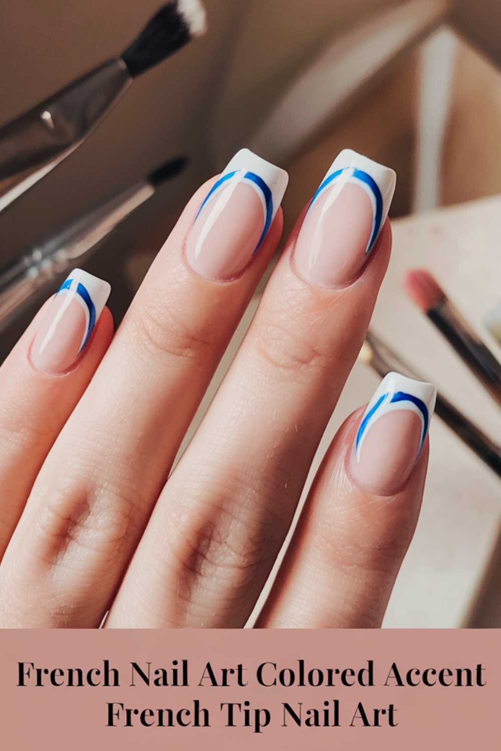 A hand with long, oval-shaped nails features a modern French manicure. The nails have white tips with a bold blue accent line beneath the tips. Various nail art brushes are blurred in the background. Text reads: French Nail Art Colored Accent French Tip Nail Art.