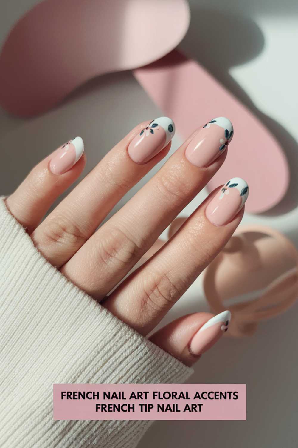 Close-up of a hand with a French manicure featuring white tips and floral accents on a pink base. The hand is partially covered by a beige sweater. The background includes soft pink elements and a faint shadow.