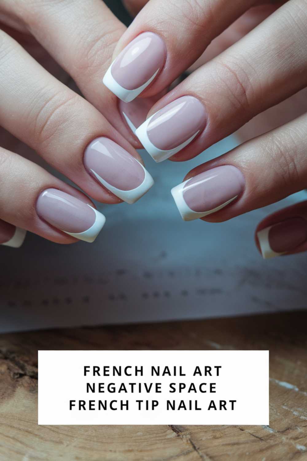 Close-up of hands with French tip nail art featuring neat white tips and nude pink polish. Text below reads, French Nail Art, Negative Space, French Tip Nail Art. Nails rest on a textured wood surface.