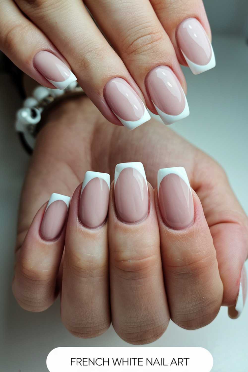 A pair of hands with a French manicure featuring glossy, nude nails with white tips. The nails are well-manicured and styled in a square shape. A bracelet is visible on one wrist, and the nails are labeled French White Nail Art.