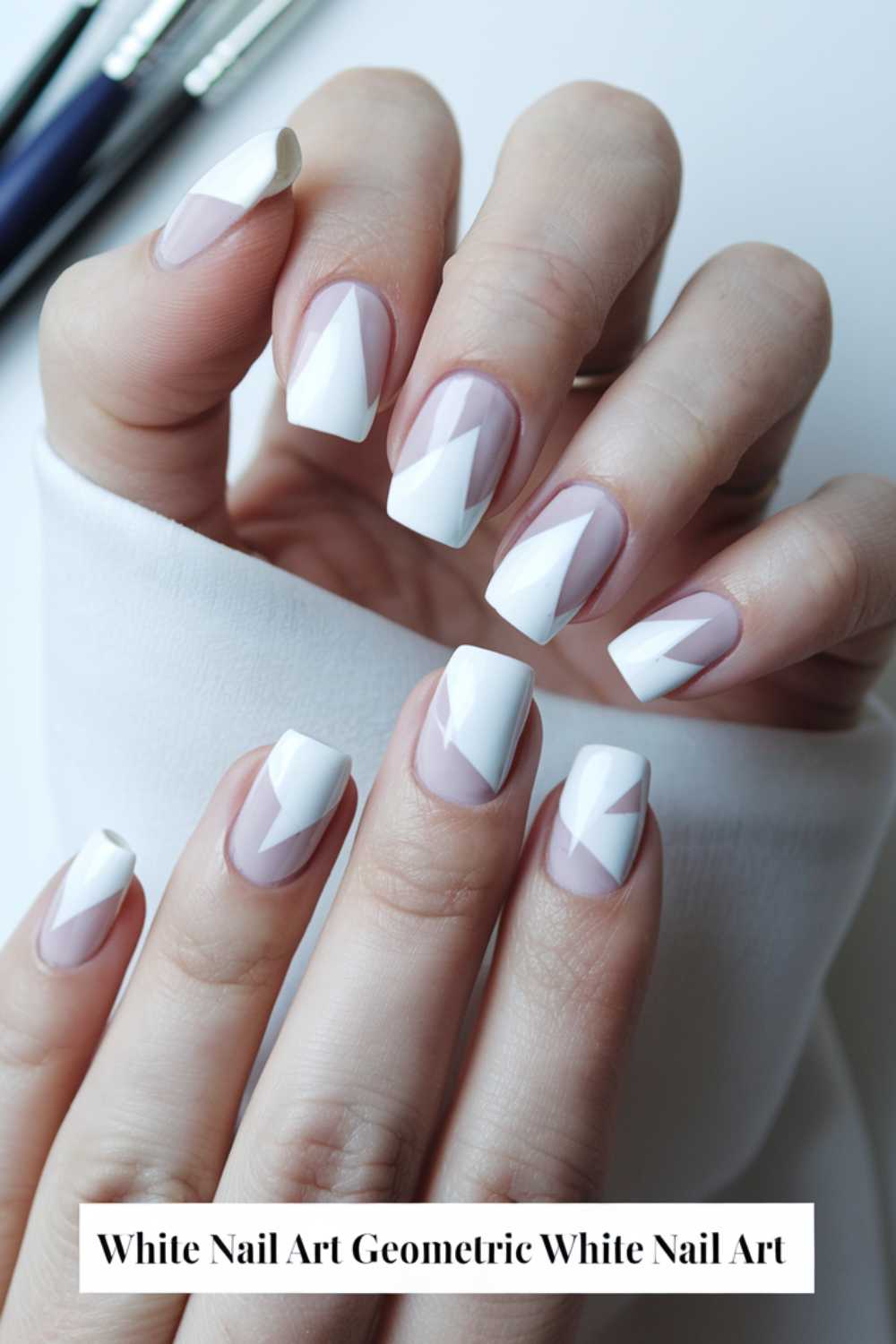 Close-up of hands with geometric white nail art. The nails feature white triangles on a neutral base, creating a modern, artistic look. The background shows part of a white garment and two pens.