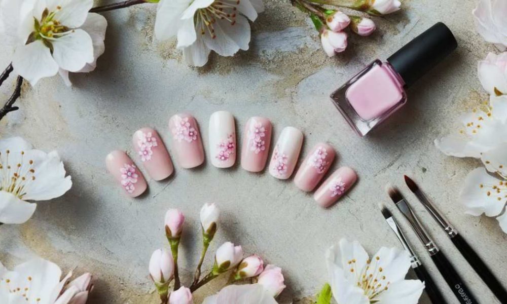 Pink floral nail art is displayed with a matching bottle of nail polish and brushes. White and pink cherry blossoms surround the scene on a textured surface.