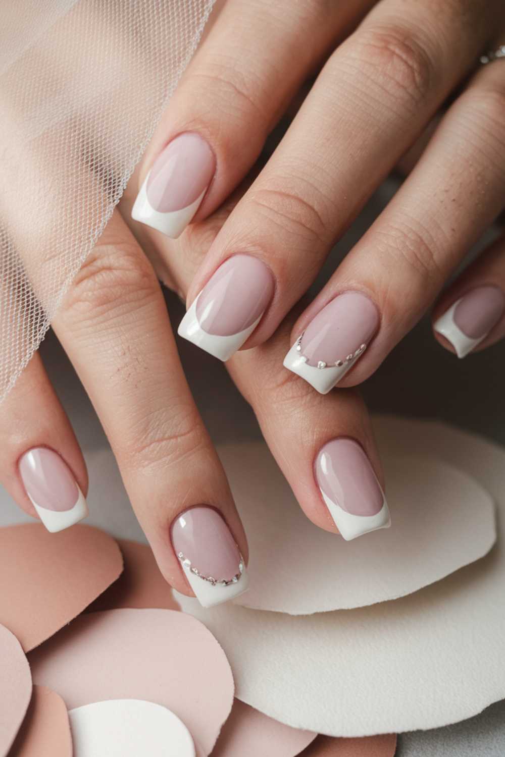 Close-up of hands with a French manicure. Nails feature pale pink polish with white tips, and one nail on each hand is adorned with small rhinestones. The fingers rest on paper cutouts in soft pink and white shades.