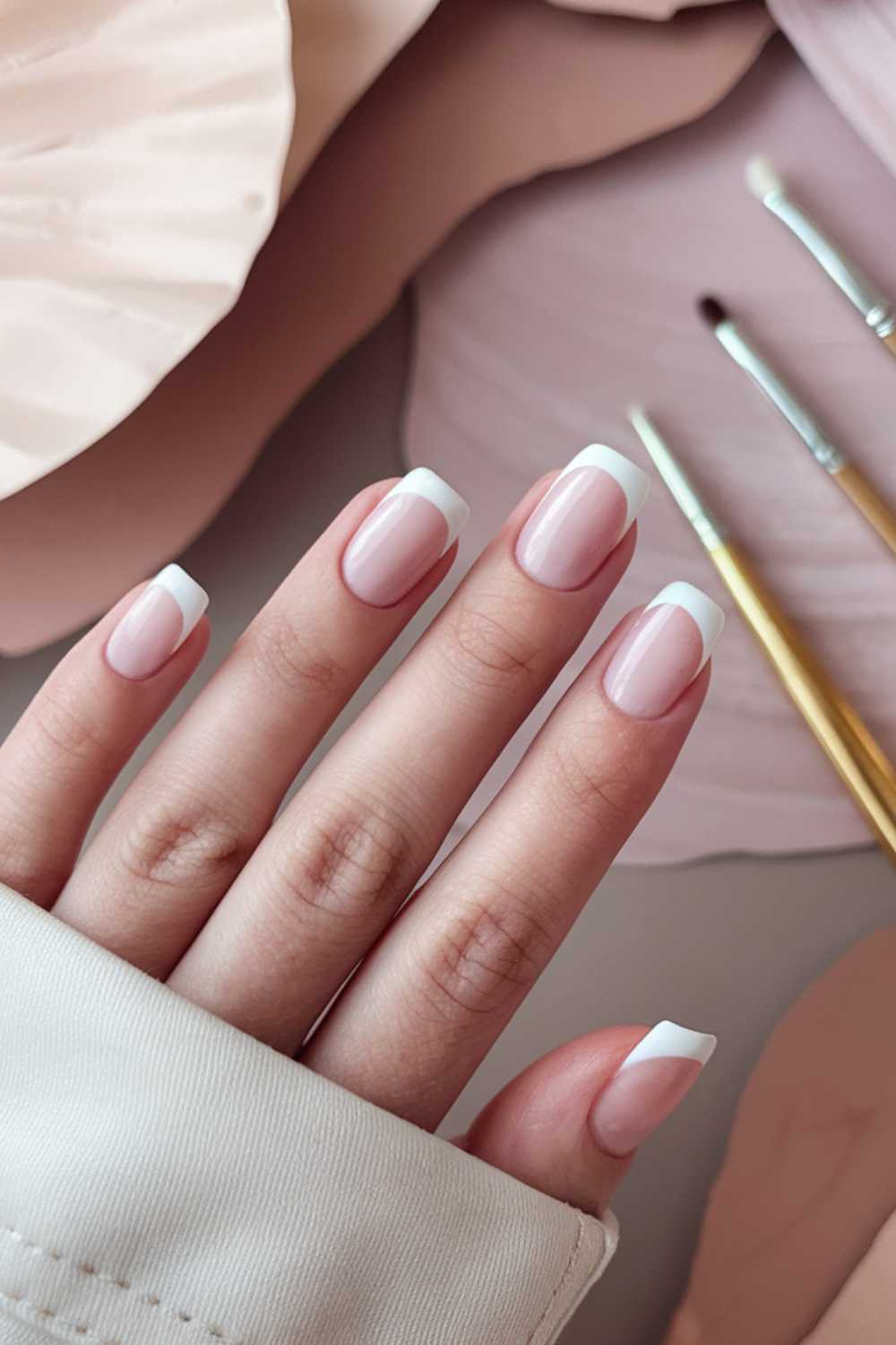 A close-up of a hand showcasing simple nail art with a French manicure against a soft beige background. The nails are neatly shaped with white tips. Two paintbrushes lie beside the hand, enhancing the aesthetic composition.