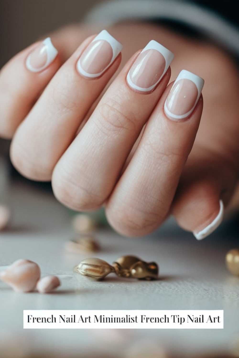 Close-up of a hand with a French manicure displaying minimalist French tip nail art. The nails have a clear base with white tips. The background features small decorative elements in soft focus.