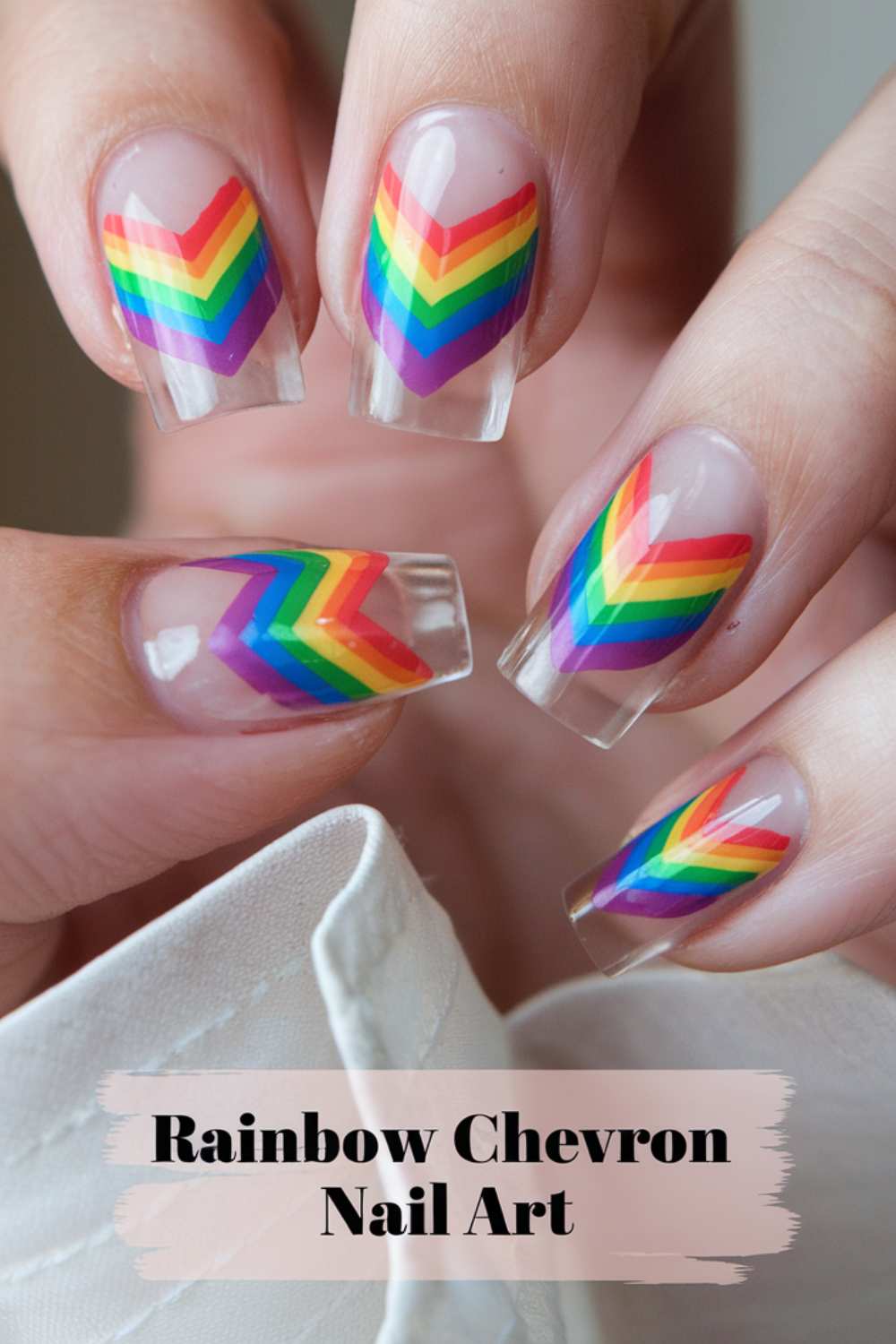 Close-up of hands displaying clear acrylic nails adorned with vibrant rainbow chevron patterns. Text at the bottom reads Rainbow Chevron Nail Art, showcasing the brilliance of rainbow nail art.