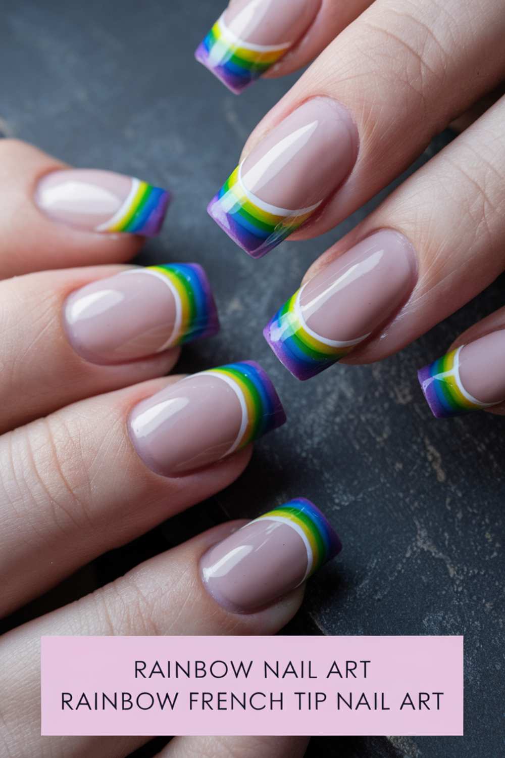 Close-up of hands with a French manicure showcasing rainbow nail art tips. The glossy nails boast vibrant stripes of purple, blue, green, yellow, and red on the tips. Below them lies a pink card that reads Rainbow Nail Art.