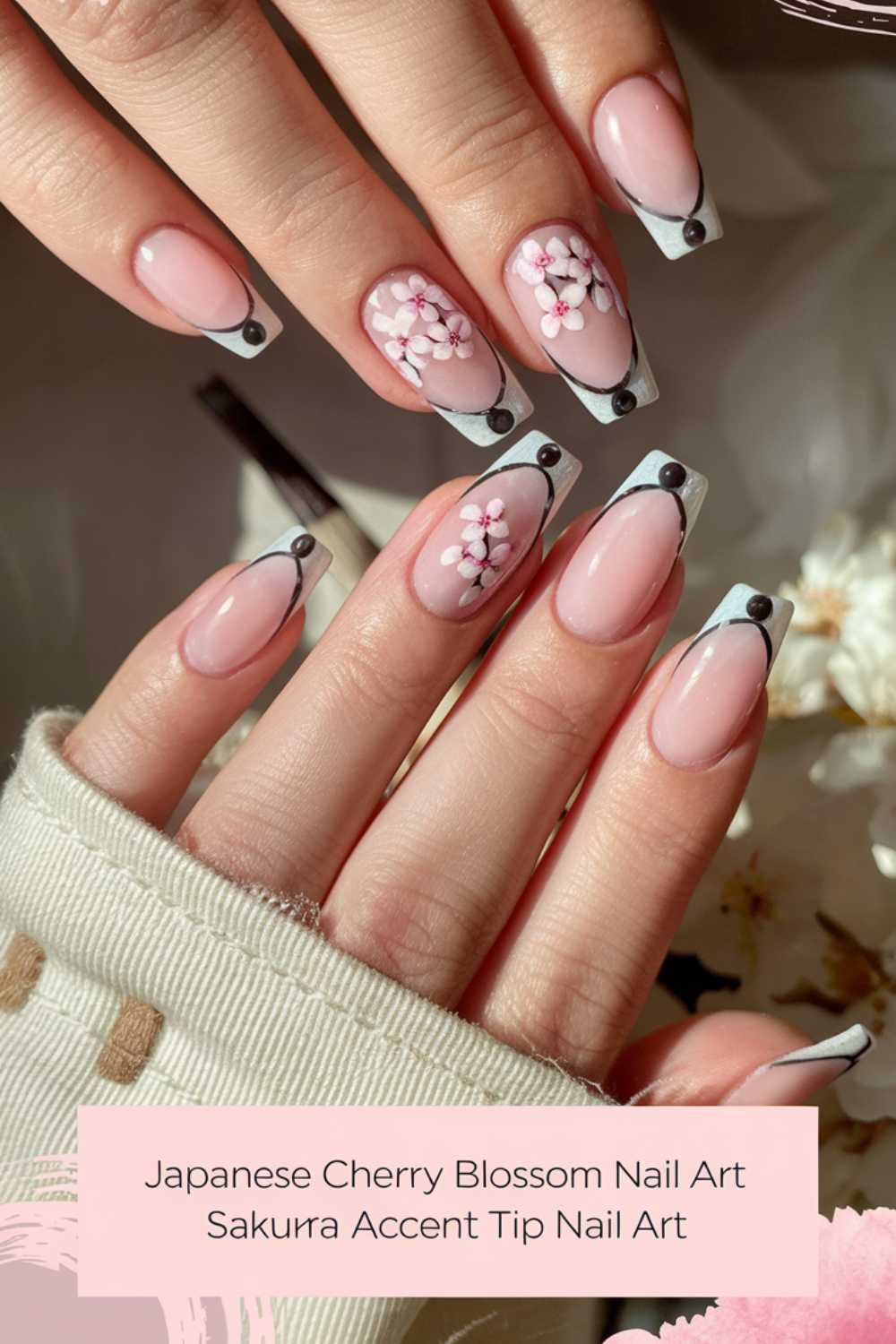 Close-up of hands with manicured nails featuring Japanese cherry blossom art. The nails have a pink base with black and white accent tips, and intricate sakura flowers are painted on some nails. The background is soft, emphasizing the nail design.