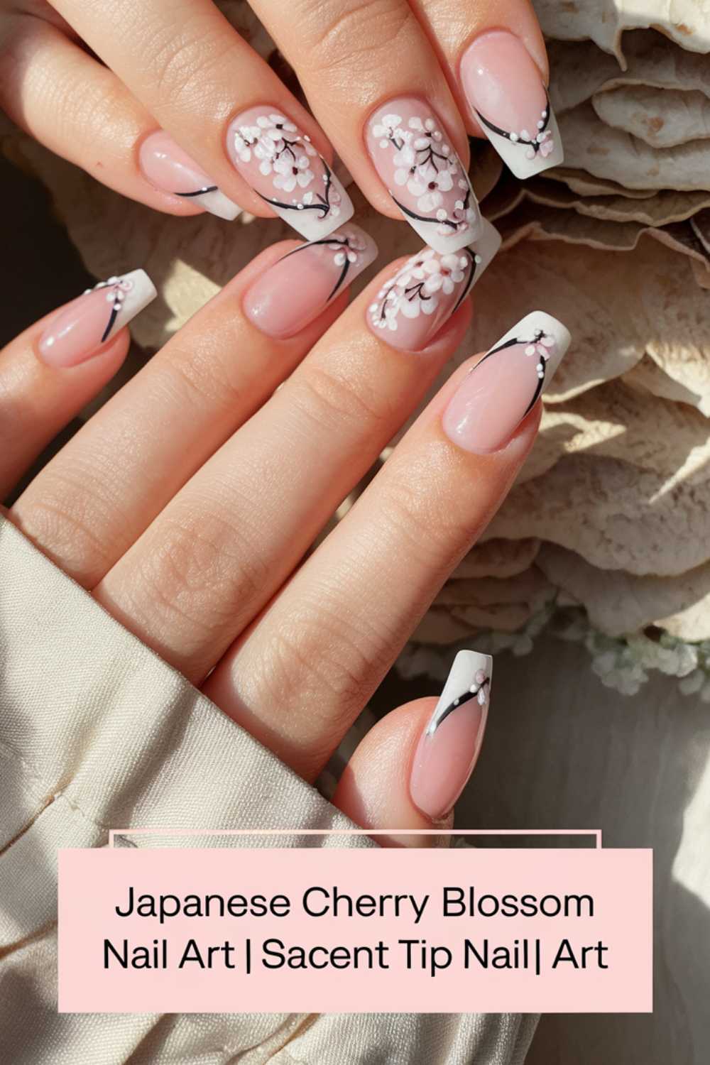 A hand displays a set of nails with a Japanese cherry blossom design. The nails are painted in soft pink with white blossoms and black branches on the tips. Text reads: Japanese Cherry Blossom Nail Art | Sacent Tip Nail | Art.
