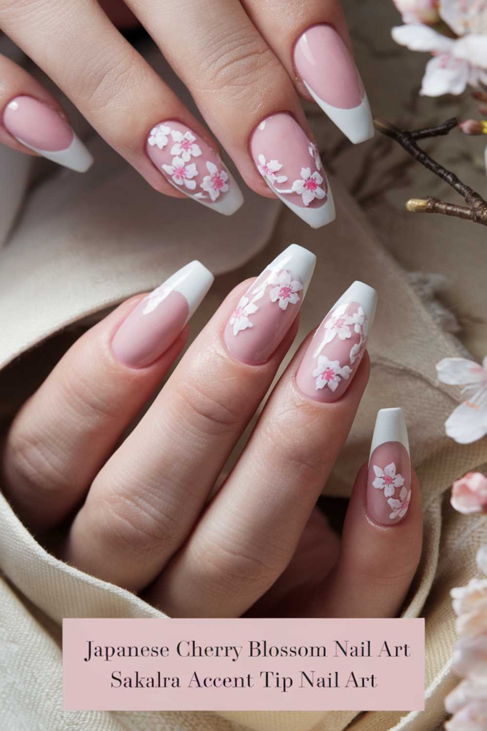 A close-up of hands with nails adorned with Japanese cherry blossom nail art. The nails feature pink and white floral designs with white tips. Nearby are cherry blossoms, complementing the elegant nail art.