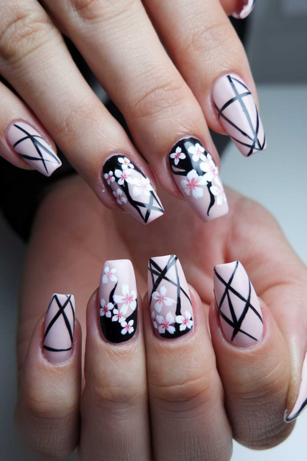 Close-up of hands showing nails with intricate designs. The nails feature pastel pink and black colors, adorned with geometric lines and small white flowers.