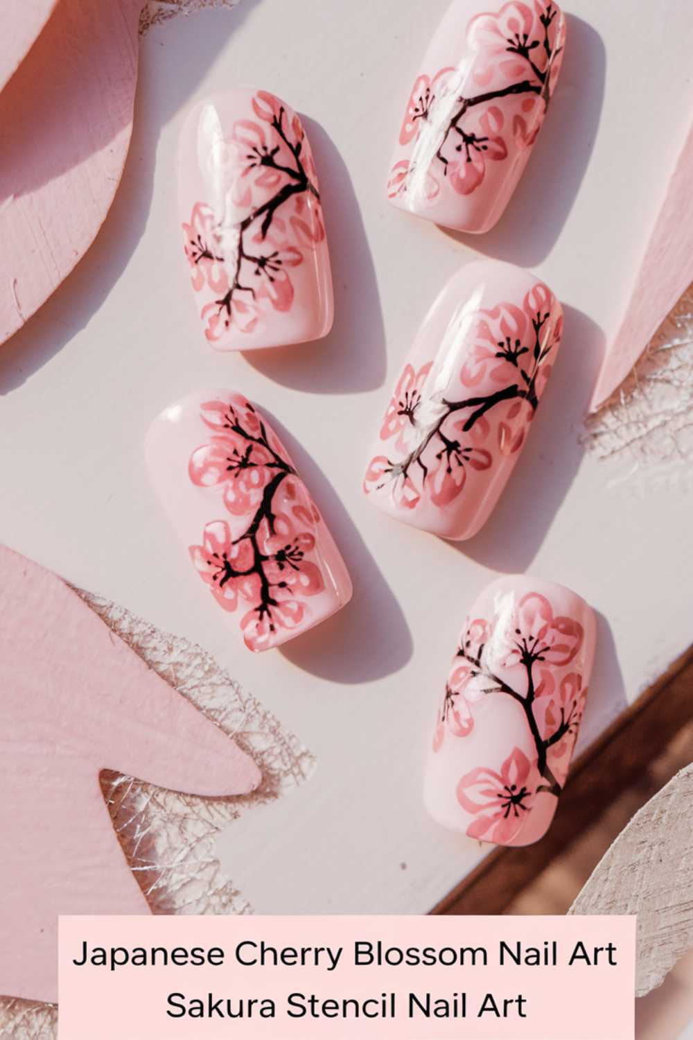 Four artificial nails with a pink base feature intricate cherry blossom designs in pink and black. The arrangement is on a textured pink surface, accompanied by the text: Japanese Cherry Blossom Nail Art Sakura Stencil Nail Art.